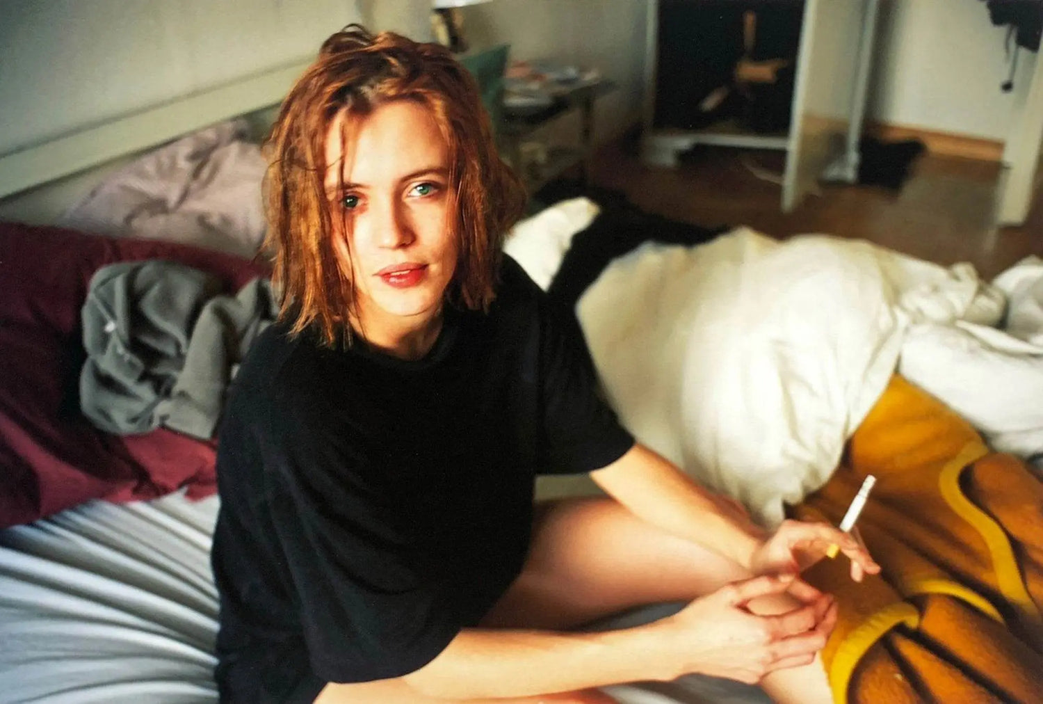 Young woman with tousled hair in black shirt sitting on bed holding a cigarette in a cozy bedroom