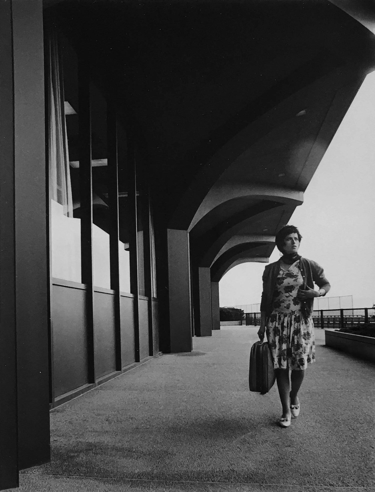 Black and white photo of woman in floral dress and cardigan walking with suitcase under modern curved building awning