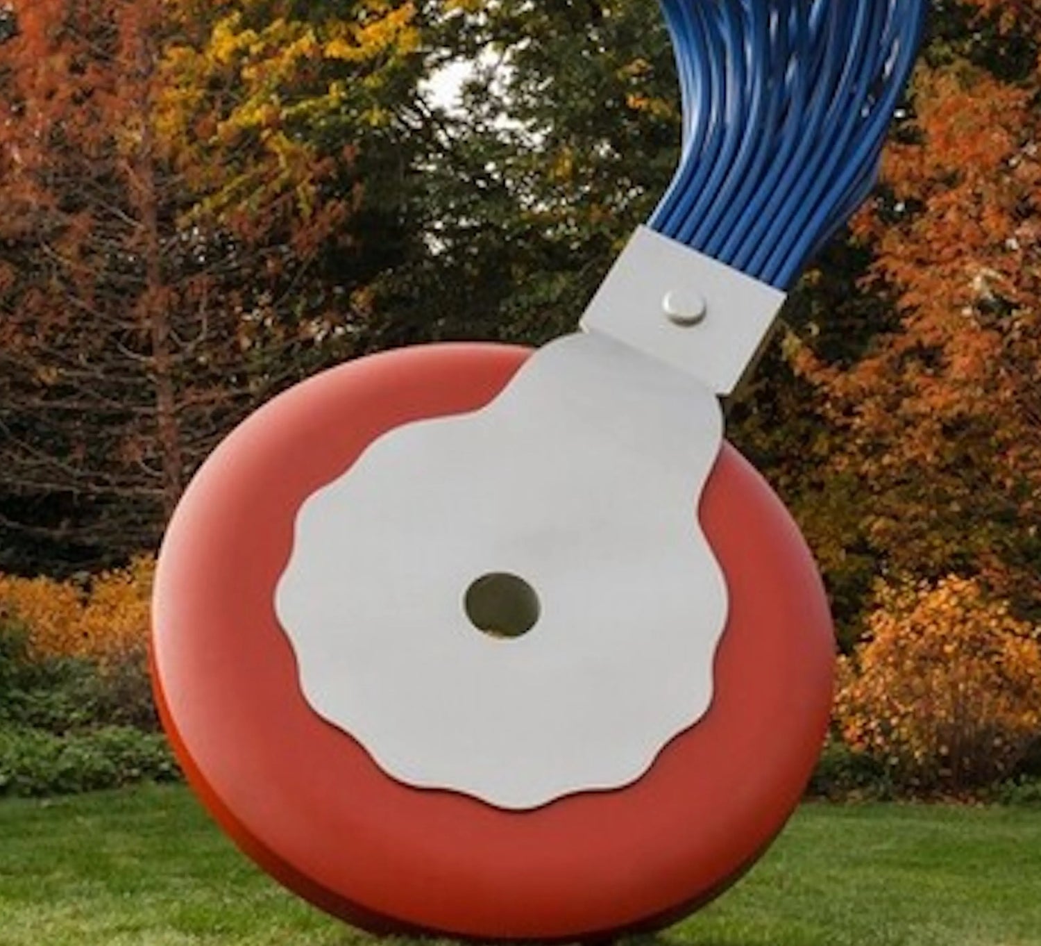 Large outdoor sculpture resembling a red and white electrical button with blue cables, set in autumn park